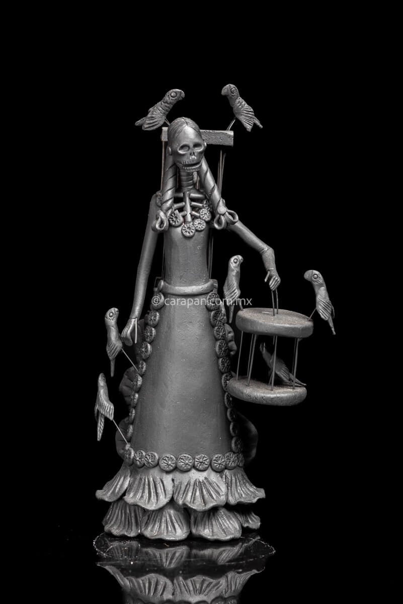 Mexican Catrina carrying a cage with parrots on her back and holding another one with her left hand. Her hair is decorated with braids and she wears a dress decorated with little rosettes on her skirt and neck.