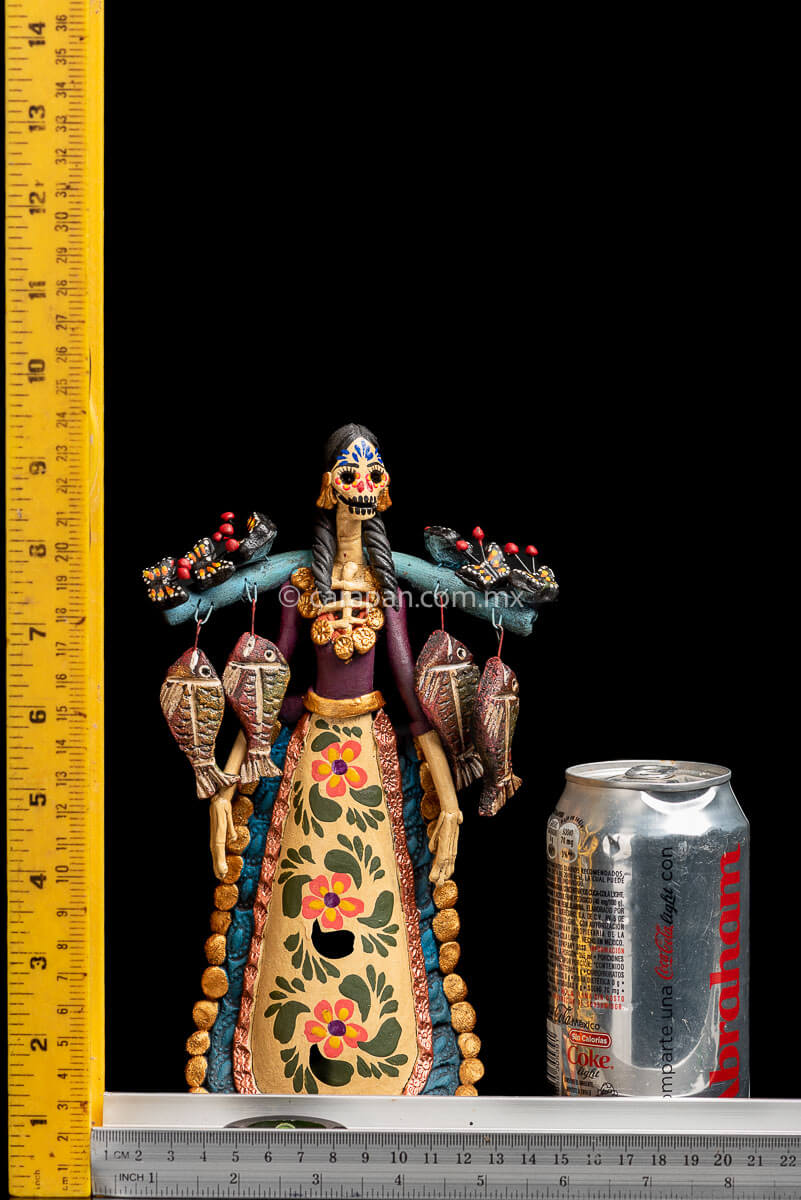 Mexican Clay Catrina carrying stick on her back with Fish and Butrerflies her skull is decorated with blue and red strokes and her dress has hand painted flowers rulers