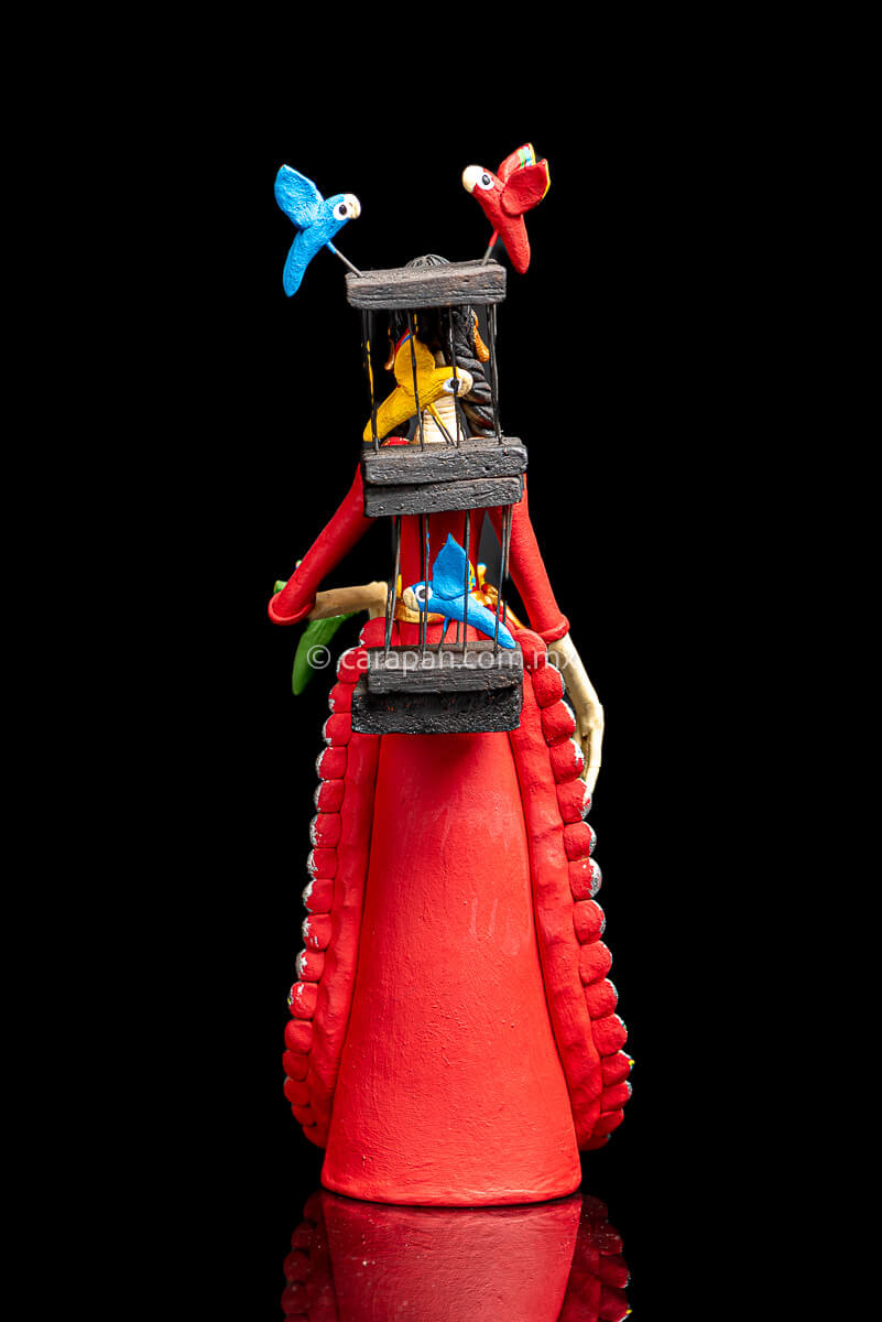 Mexican Clay Catrina holding a bird cage with her left hand and carrying another on her back her skull is painted with strokes in green, red yellow and blue. Her skirt is painted with purple flowers and green leafs red back