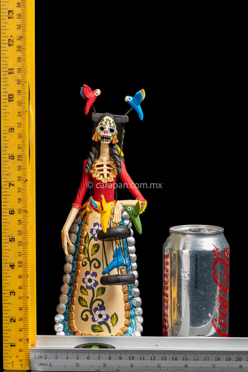 Mexican Clay Catrina holding a bird cage with her left hand and carrying another on her back her skull is painted with strokes in green, red yellow and blue. Her skirt is painted with purple flowers and green leafs rulers