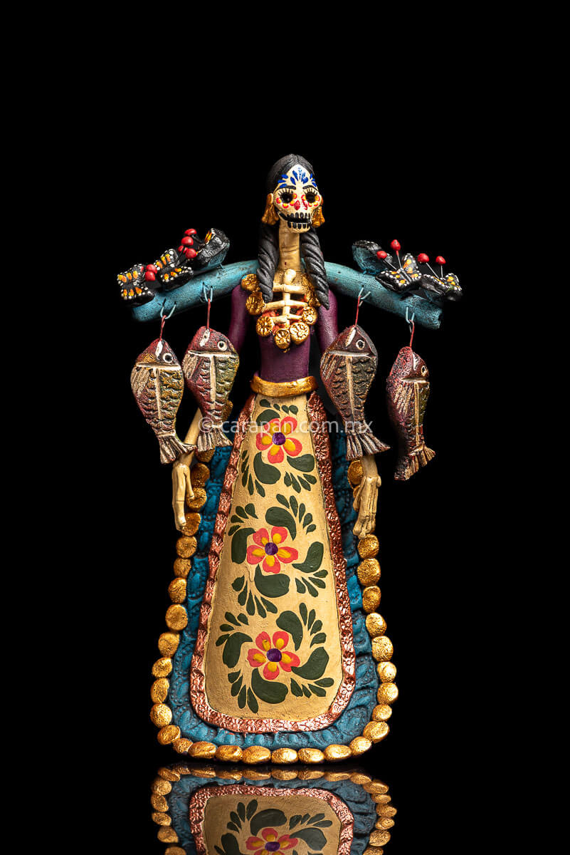 Mexican Clay Catrina carrying stick on her back with Fish and Butrerflies her skull is decorated with blue and red strokes and her dress has hand painted flowers
