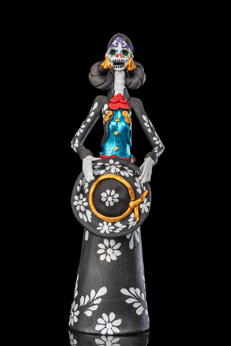 Catrina day of the dead Clay Sculpture dressed in a black and white charro dress  holding her hat on  her waist  with both hands.  Her skull is  hand painted.