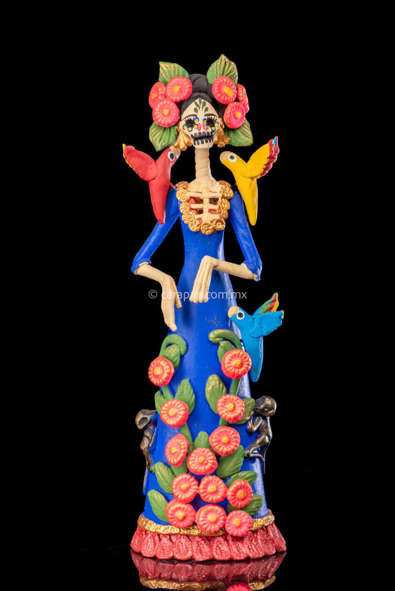 Mexican Clay Catrina Sculpture with blue dress and pink flowers. The catrina is inspired by Two of the most emblematic Frida Kahlo's self portraits with monkeys and parrots. Taht's why this catrina has three parrots and monkeys next to her. Her head dress is decorated with pink flowers and her skull is hand painted.