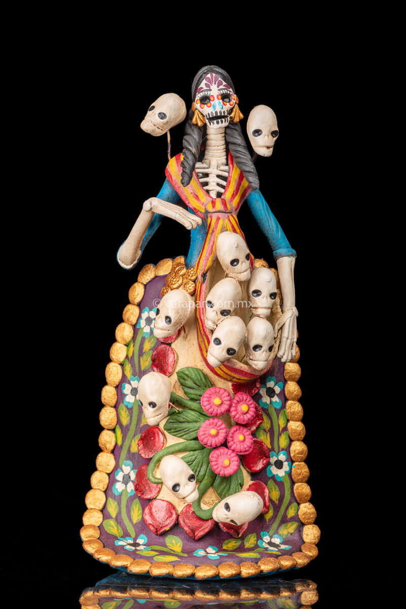 Catrina clay sculpture wearing a traditional Purépecha dress with a rebozo.  She carries 5 skulls in her rebozo and 4 more skulls adorn her dress. Two more skulls are next to her above each of her shoulders. Her dress is also decorated with pastillage flowers and painted flowers. Her own skull is hand painted in vibrant colors as well as her dress