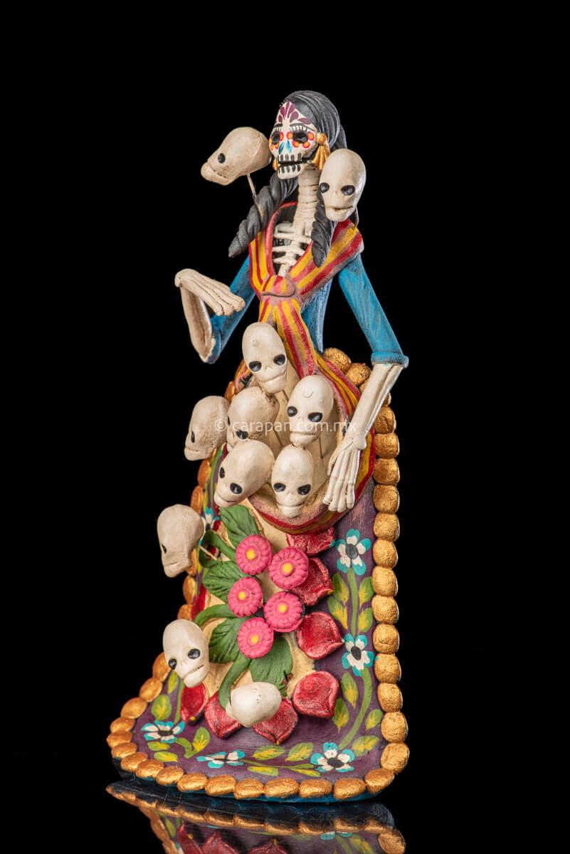 Catrina clay sculpture wearing a traditional Purépecha dress with a rebozo.  She carries 5 skulls in her rebozo and 4 more skulls adorn her dress. Two more skulls are next to her above each of her shoulders. Her dress is also decorated with pastillage flowers and painted flowers. Her own skull is hand painted in vibrant colors as well as her dress