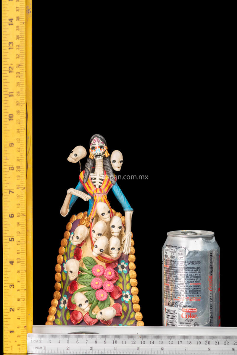Catrina clay sculpture wearing a traditional Purépecha dress with a rebozo.  She carries 5 skulls in her rebozo and 4 more skulls adorn her dress. Two more skulls are next to her above each of her shoulders. Her dress is also decorated with pastillage flowers and painted flowers. Her own skull is hand painted in vibrant colors as well as her dress