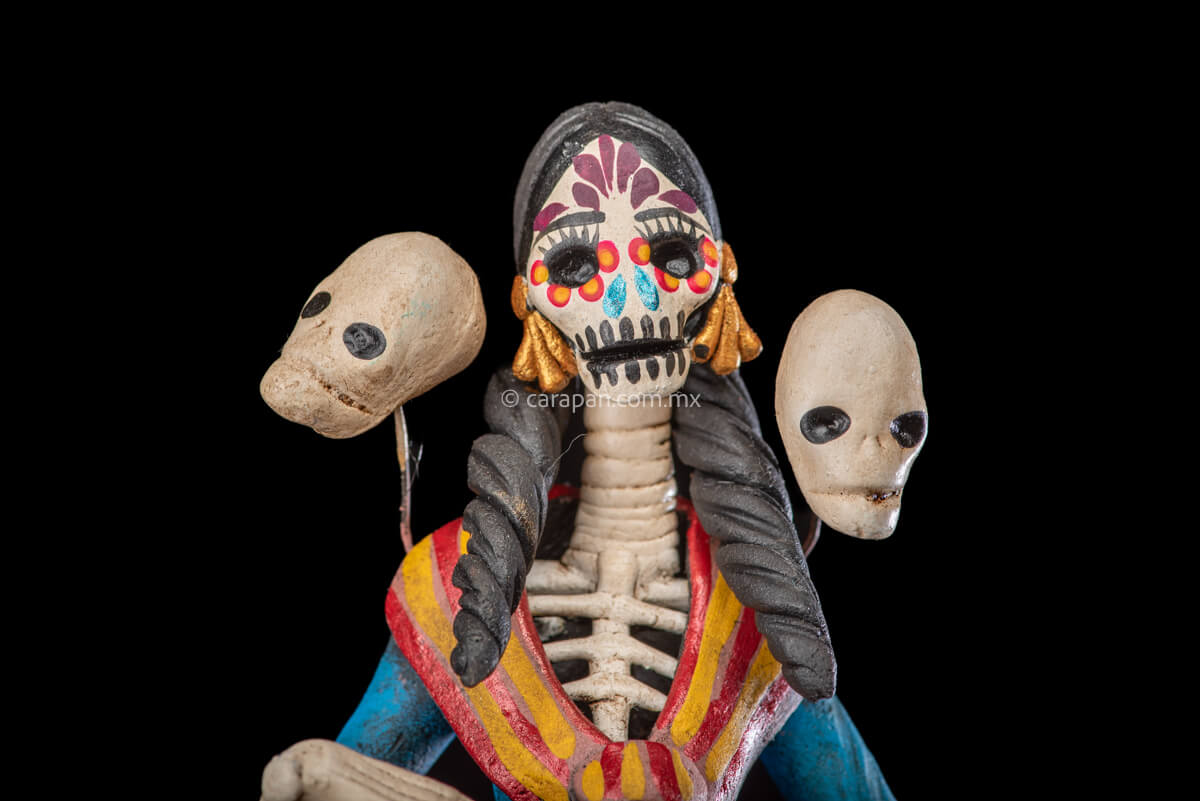 Catrina clay sculpture wearing a traditional Purépecha dress with a rebozo.  She carries 5 skulls in her rebozo and 4 more skulls adorn her dress. Two more skulls are next to her above each of her shoulders. Her dress is also decorated with pastillage flowers and painted flowers. Her own skull is hand painted in vibrant colors as well as her dress