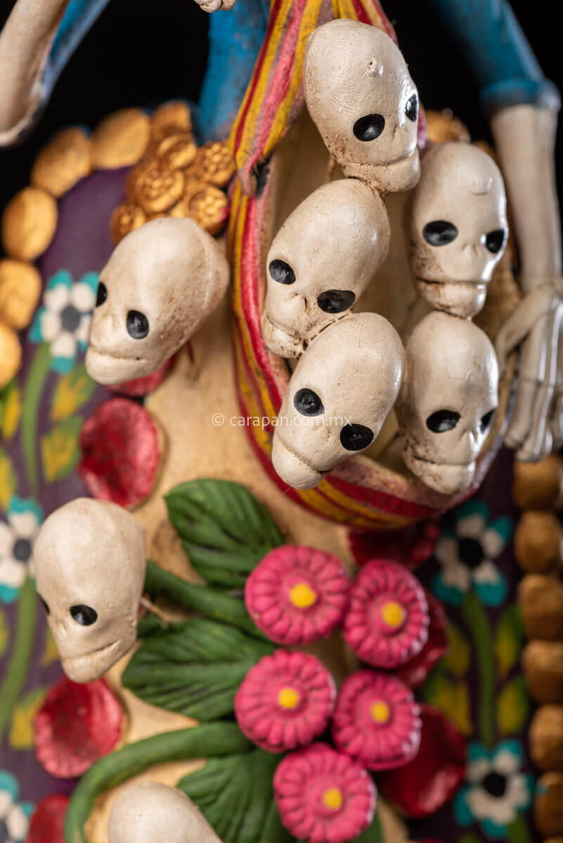 Catrina clay sculpture wearing a traditional Purépecha dress with a rebozo.  She carries 5 skulls in her rebozo and 4 more skulls adorn her dress. Two more skulls are next to her above each of her shoulders. Her dress is also decorated with pastillage flowers and painted flowers. Her own skull is hand painted in vibrant colors as well as her dress