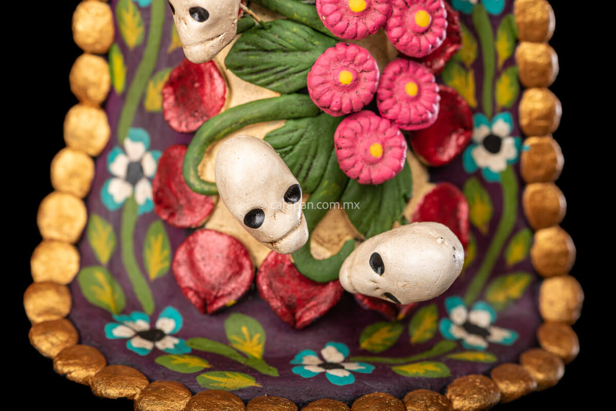 Catrina clay sculpture wearing a traditional Purépecha dress with a rebozo.  She carries 5 skulls in her rebozo and 4 more skulls adorn her dress. Two more skulls are next to her above each of her shoulders. Her dress is also decorated with pastillage flowers and painted flowers. Her own skull is hand painted in vibrant colors as well as her dress