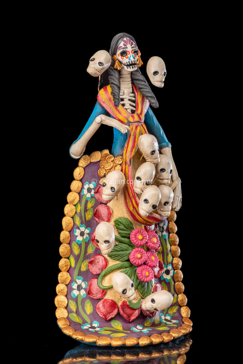 Catrina clay sculpture wearing a traditional Purépecha dress with a rebozo.  She carries 5 skulls in her rebozo and 4 more skulls adorn her dress. Two more skulls are next to her above each of her shoulders. Her dress is also decorated with pastillage flowers and painted flowers. Her own skull is hand painted in vibrant colors as well as her dress
