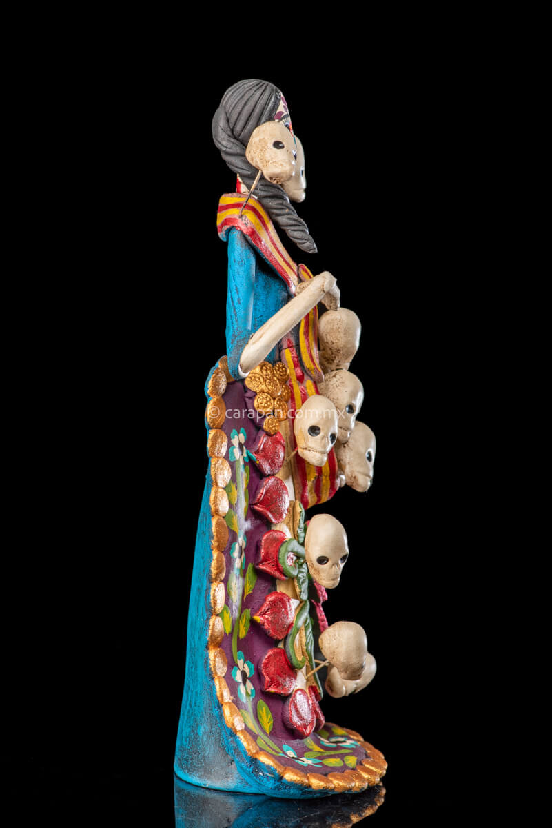 Catrina clay sculpture wearing a traditional Purépecha dress with a rebozo.  She carries 5 skulls in her rebozo and 4 more skulls adorn her dress. Two more skulls are next to her above each of her shoulders. Her dress is also decorated with pastillage flowers and painted flowers. Her own skull is hand painted in vibrant colors as well as her dress
