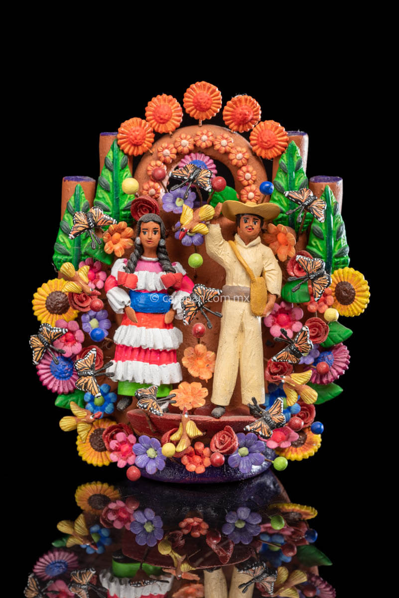 Small Mexican Clay tree of life with Mazahua indigenous couple at the center surrounded by flowers and butterflies. The man wears a traditional costume in beig, hat and a bag. The woman wears a traditional dress in pink, orange and green with a blue belt.