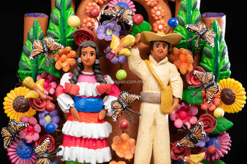 Small Mexican Clay tree of life with Mazahua indigenous couple at the center surrounded by flowers and butterflies. The man wears a traditional costume in beig, hat and a bag. The woman wears a traditional dress in pink, orange and green with a blue belt.