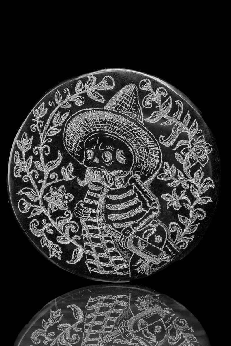 Skeleton  wearing a sombrero, a sarape and holding a bottle based upon a Jose Guadalupe Posadas drawing. The piece is framed by flowers and vegetal motifs.