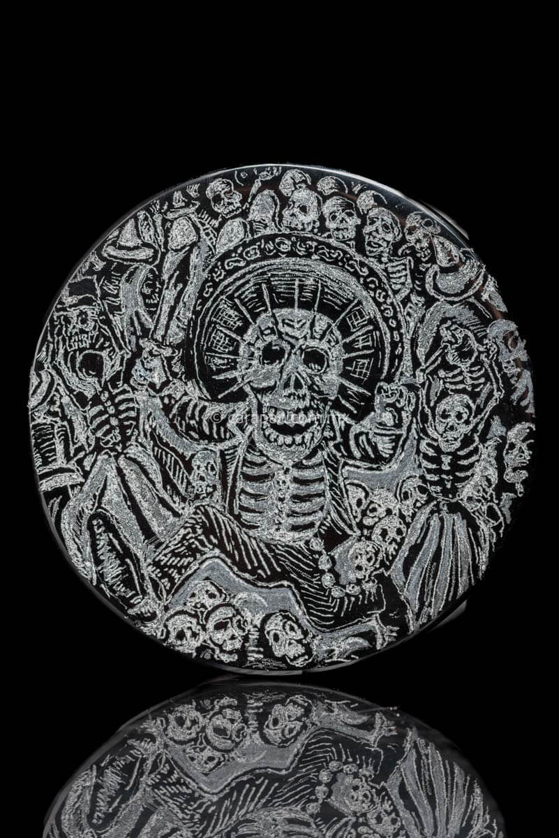 Obsidian disc with engraved skeleton wearing a charro costume, holding a knife on his right hand and running . Surrounded by skulls and skeleton. Based on Jose Guadalupe Posadas Work