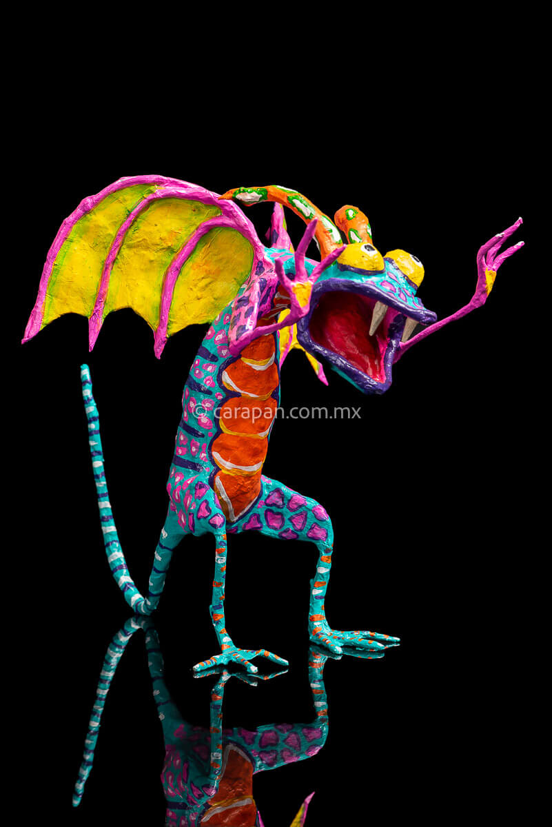 Paper Mache Alebrije Dragon Style Standing with hands up and mouth open showing 2 fangs popping yellow eyes and two orange horns. His body is painted in turquoise with pink dots and blue stripes and his wings are yellow outlined in pink
