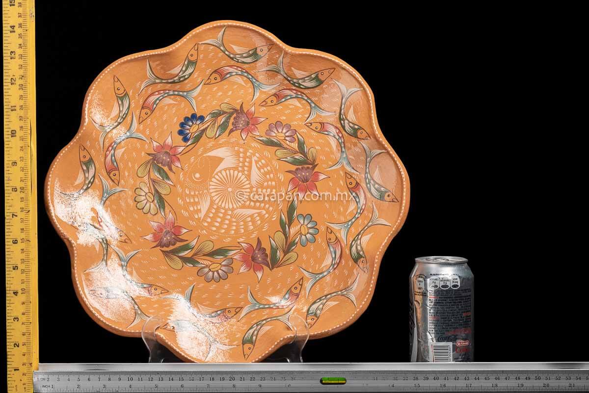 Light orange dish with curvy flower shape decorated with two circles of fishes painted in green and dark orange following by a circle of flowers and one  fish in beige at the center with its mouth near its tail in circular position. Finally, a star in beige with a white dot and strokesat the center rulers