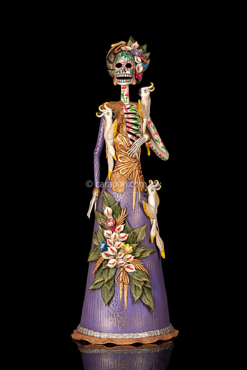Mexican Catrina, Day of the dead Sculpture wearing a purple dress with pastillage calla lilies at the bottom. The skull's face is painted and has a head dress with calla lilies three parrots are attached to her body. One near her left shoulder, one  near her right chest and one at her left side of her waist 