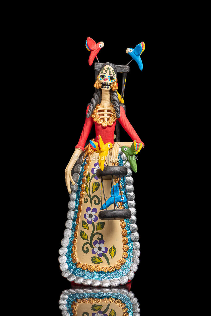 Mexican Clay Catrina holding a bird cage with her left hand and carrying another on her back her skull is painted with strokes in green, red yellow and blue. Her skirt is painted with purple flowers and green leafs