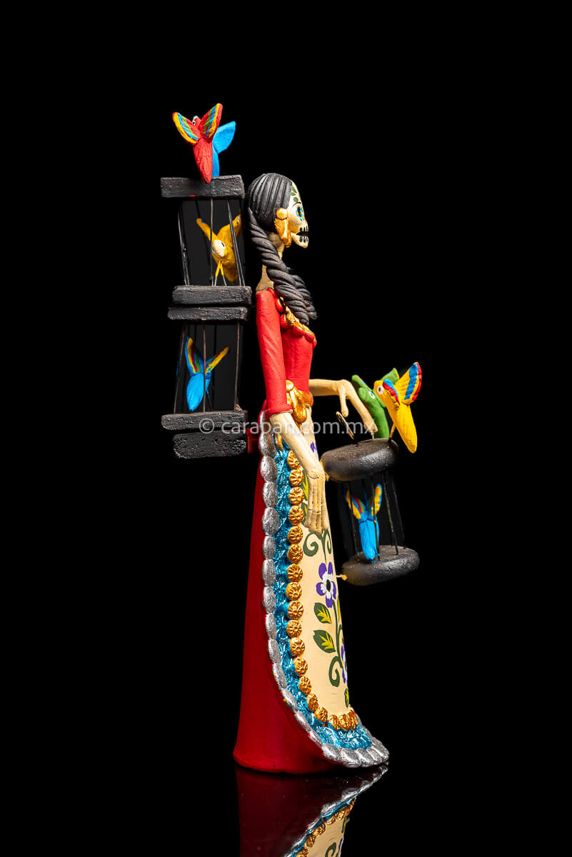 Mexican Clay Catrina holding a bird cage with her left hand and carrying another on her back her skull is painted with strokes in green, red yellow and blue. Her skirt is painted with purple flowers and green leafs Side 1