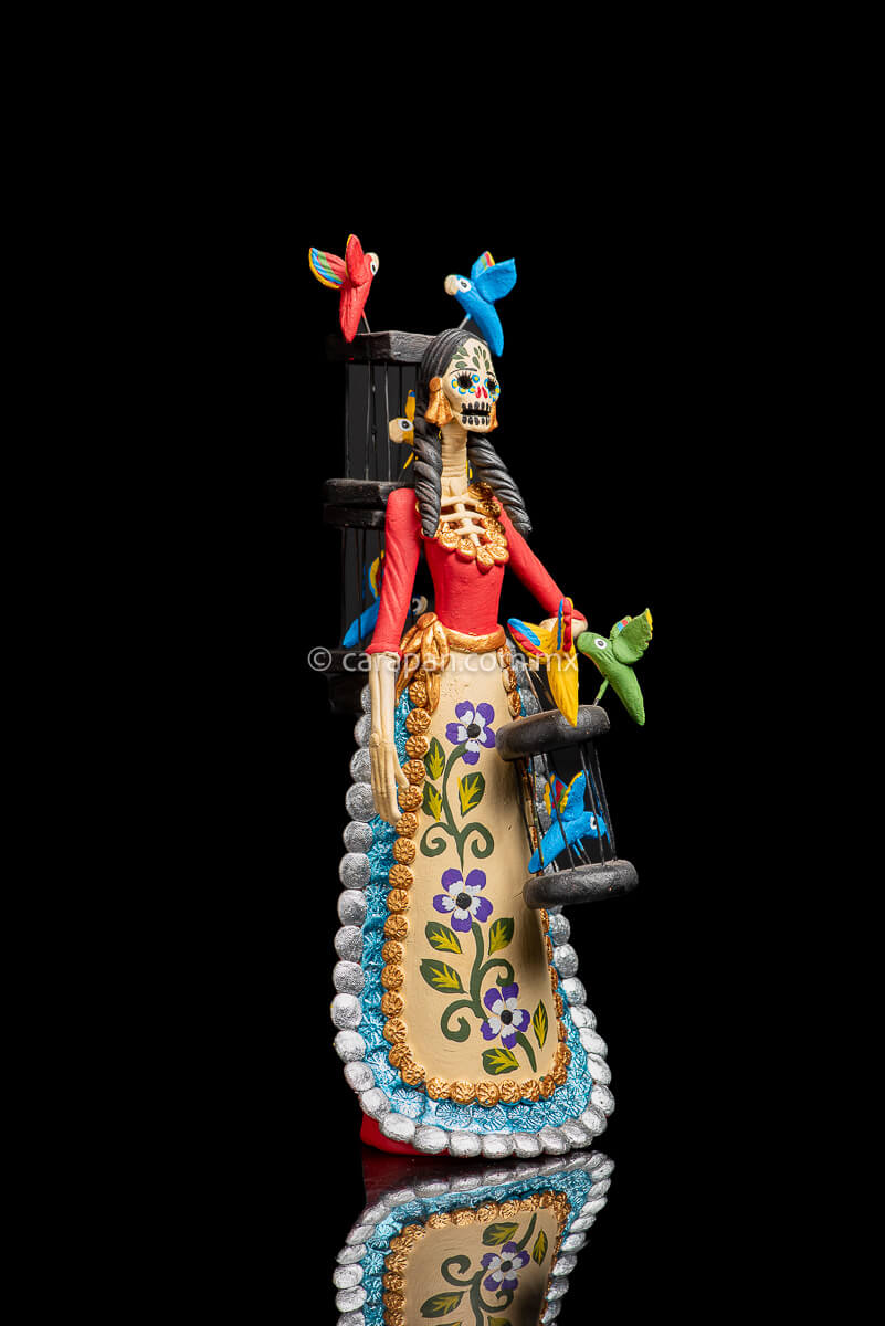 Mexican Clay Catrina holding a bird cage with her left hand and carrying another on her back her skull is painted with strokes in green, red yellow and blue. Her skirt is painted with purple flowers and green leafs facing right