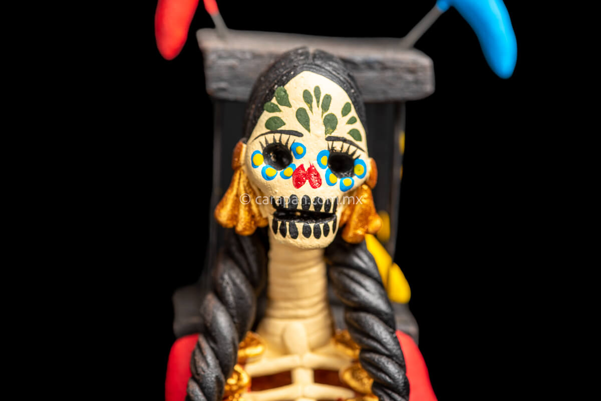 Mexican Clay Catrina holding a bird cage with her left hand and carrying another on her back her skull is painted with strokes in green, red yellow and blue. Her skirt is painted with purple flowers and green leafs Skull close up