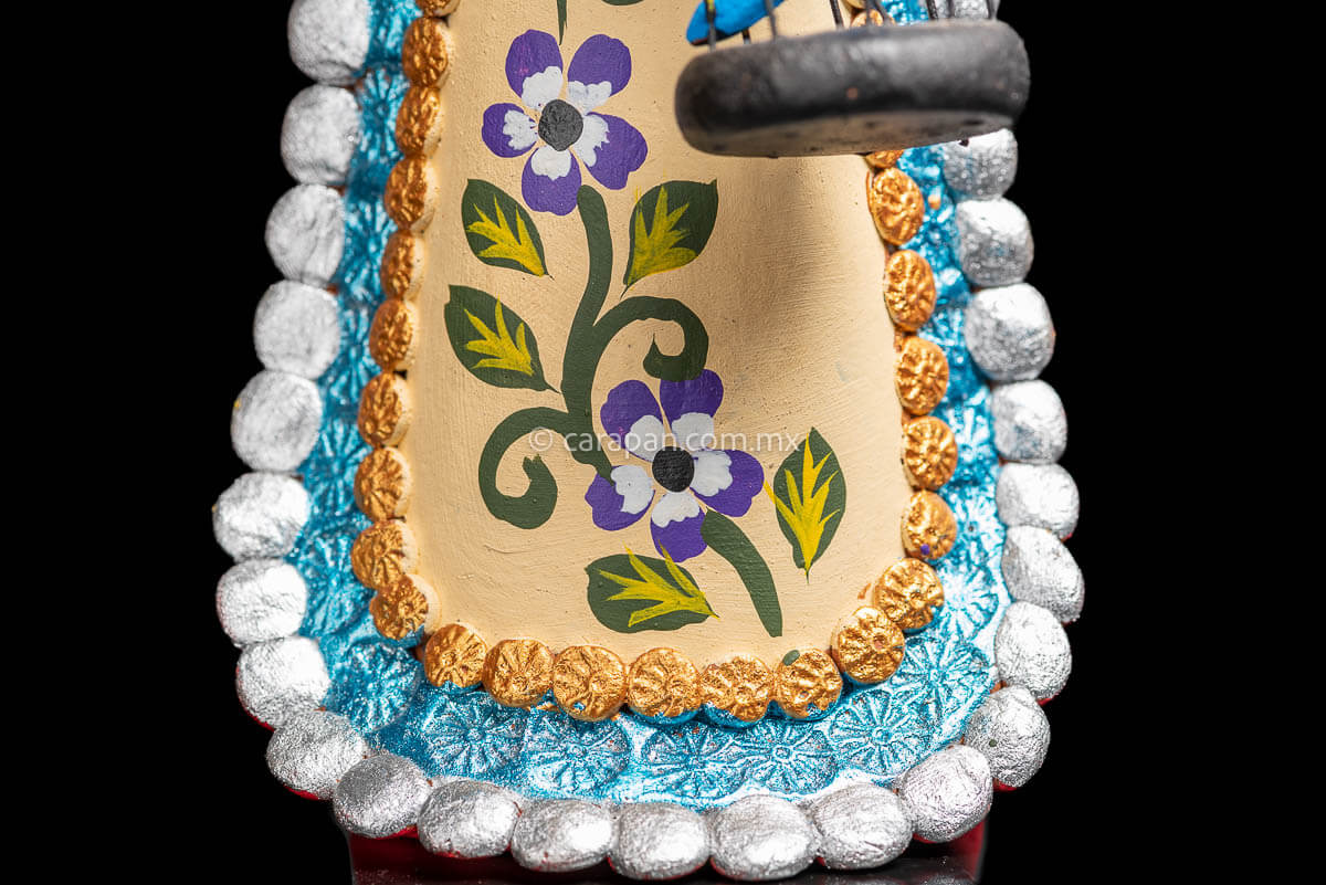 Mexican Clay Catrina holding a bird cage with her left hand and carrying another on her back her skull is painted with strokes in green, red yellow and blue. Her skirt is painted with purple flowers and green leafs Skirt detail