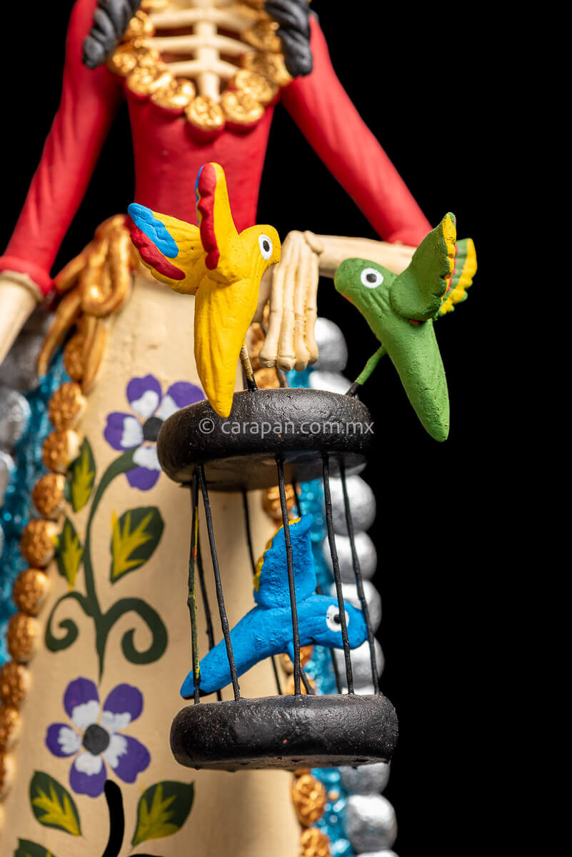 Mexican Clay Catrina holding a bird cage with her left hand and carrying another on her back her skull is painted with strokes in green, red yellow and blue. Her skirt is painted with purple flowers and green leafs Birds