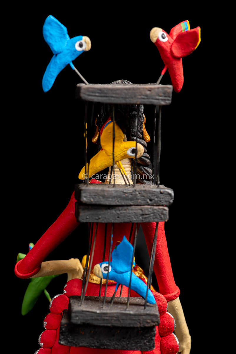 Mexican Clay Catrina holding a bird cage with her left hand and carrying another on her back her skull is painted with strokes in green, red yellow and blue. Her skirt is painted with purple flowers and green leafs bird cages