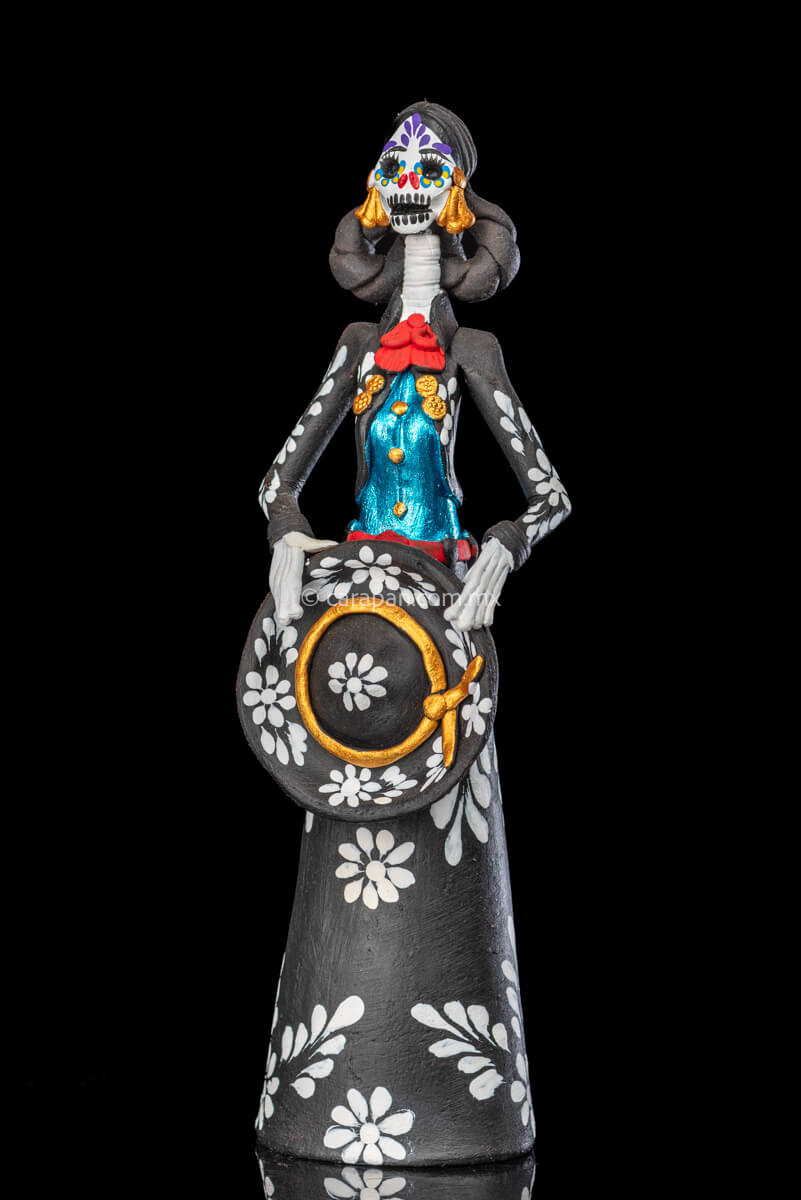 Catrina day of the dead Clay Sculpture dressed in a black and white charro dress  holding her hat on  her waist  with both hands.  Her skull is  hand painted. 