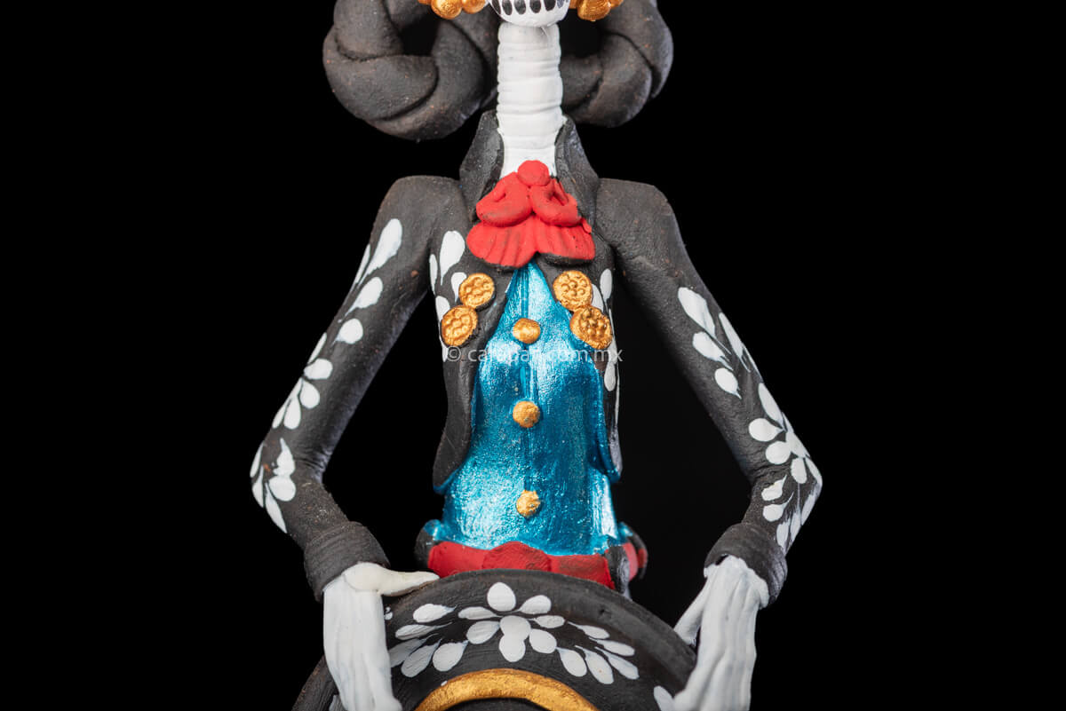 Catrina day of the dead Clay Sculpture dressed in a black and white charro dress  holding her hat on  her waist  with both hands.  Her skull is  hand painted.  detail