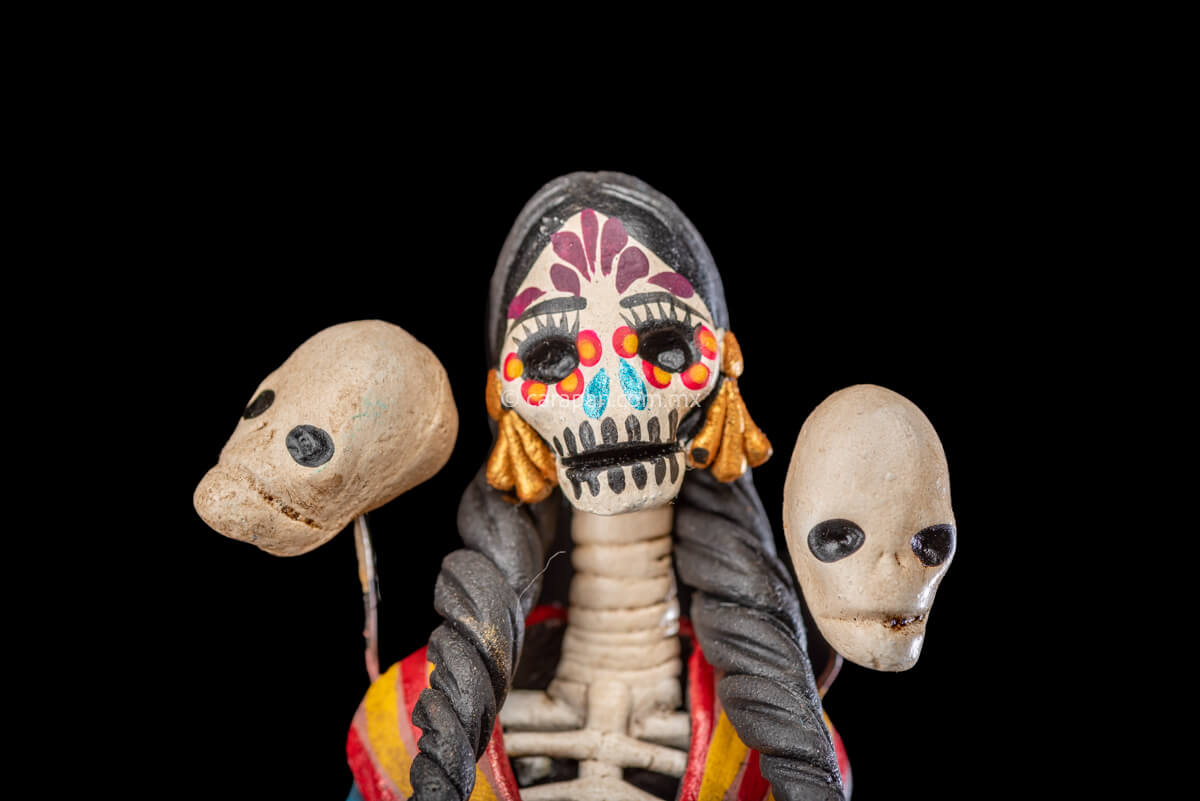 Catrina clay sculpture wearing a traditional Purépecha dress with a rebozo.  She carries 5 skulls in her rebozo and 4 more skulls adorn her dress. Two more skulls are next to her above each of her shoulders. Her dress is also decorated with pastillage flowers and painted flowers. Her own skull is hand painted in vibrant colors as well as her dress