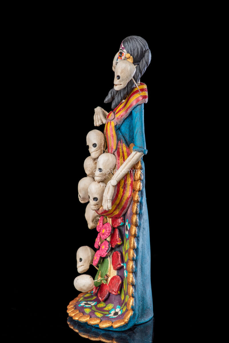 Catrina clay sculpture wearing a traditional Purépecha dress with a rebozo.  She carries 5 skulls in her rebozo and 4 more skulls adorn her dress. Two more skulls are next to her above each of her shoulders. Her dress is also decorated with pastillage flowers and painted flowers. Her own skull is hand painted in vibrant colors as well as her dress