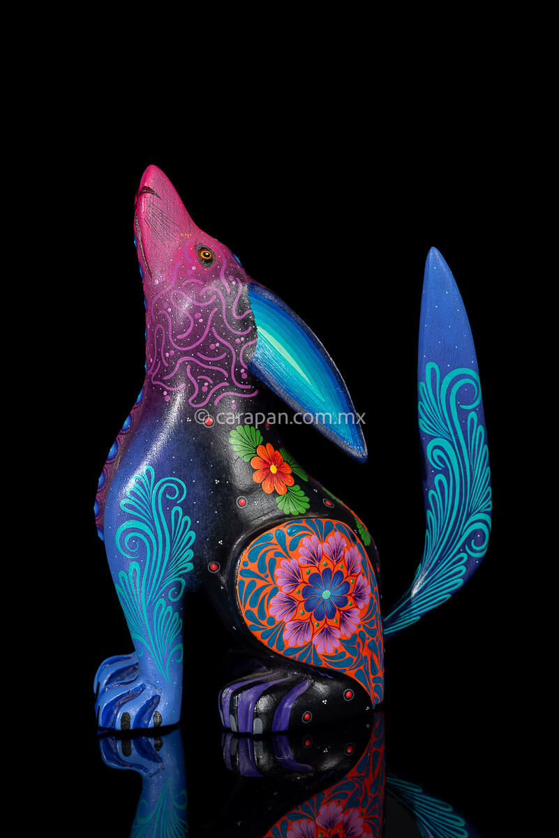 Wood carving of a Coyote looking up its body has blue, black and purple hues and is decorated with flowers and dots that resemble the stars