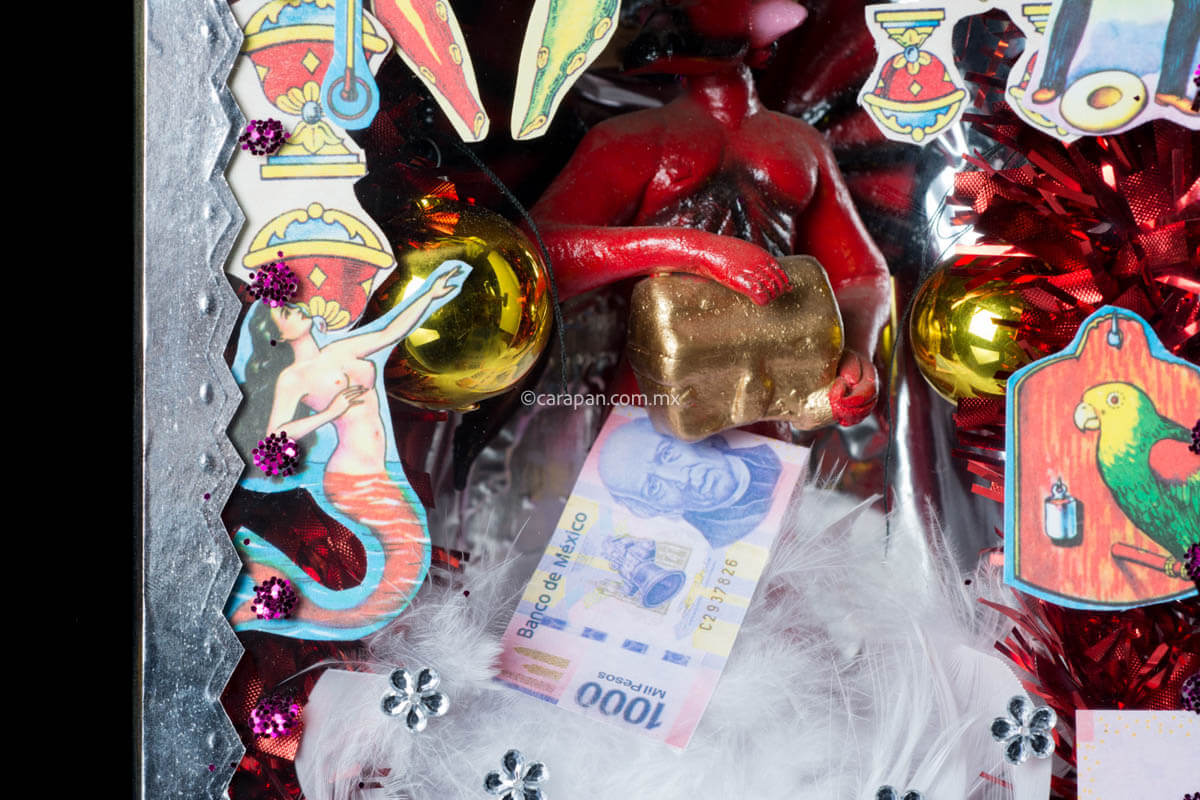 Diorama with Clay Devil holding a golden box surrouded by Chrsitmas spheres and white feathers. the glass of the diorama is decorated with clippings of Spanish Cards and Mexican Lottery. The top of the diorama is decorated with shiny flowers in gold and silver and has a clipping of a shampoo ad with a woman.