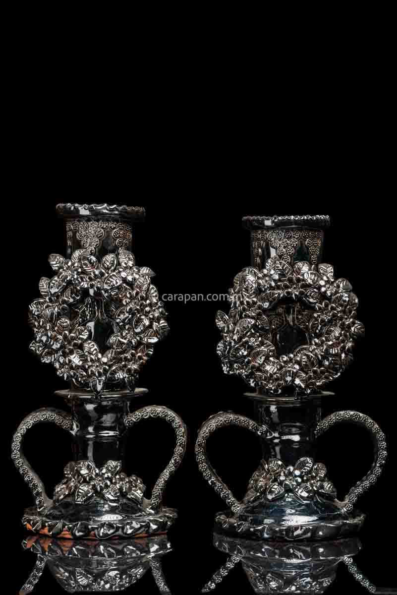 Set of 2 black glazed candlesticks. Each is decorated with a profuse pastillage crown of flowers. At the center of each crown is a couple of loving birds. Each candlestick has two  curved handles at the bottom. The contour of the handles is decorated with rosettes and the base of each candlestick is decorated with flowers and leafs.