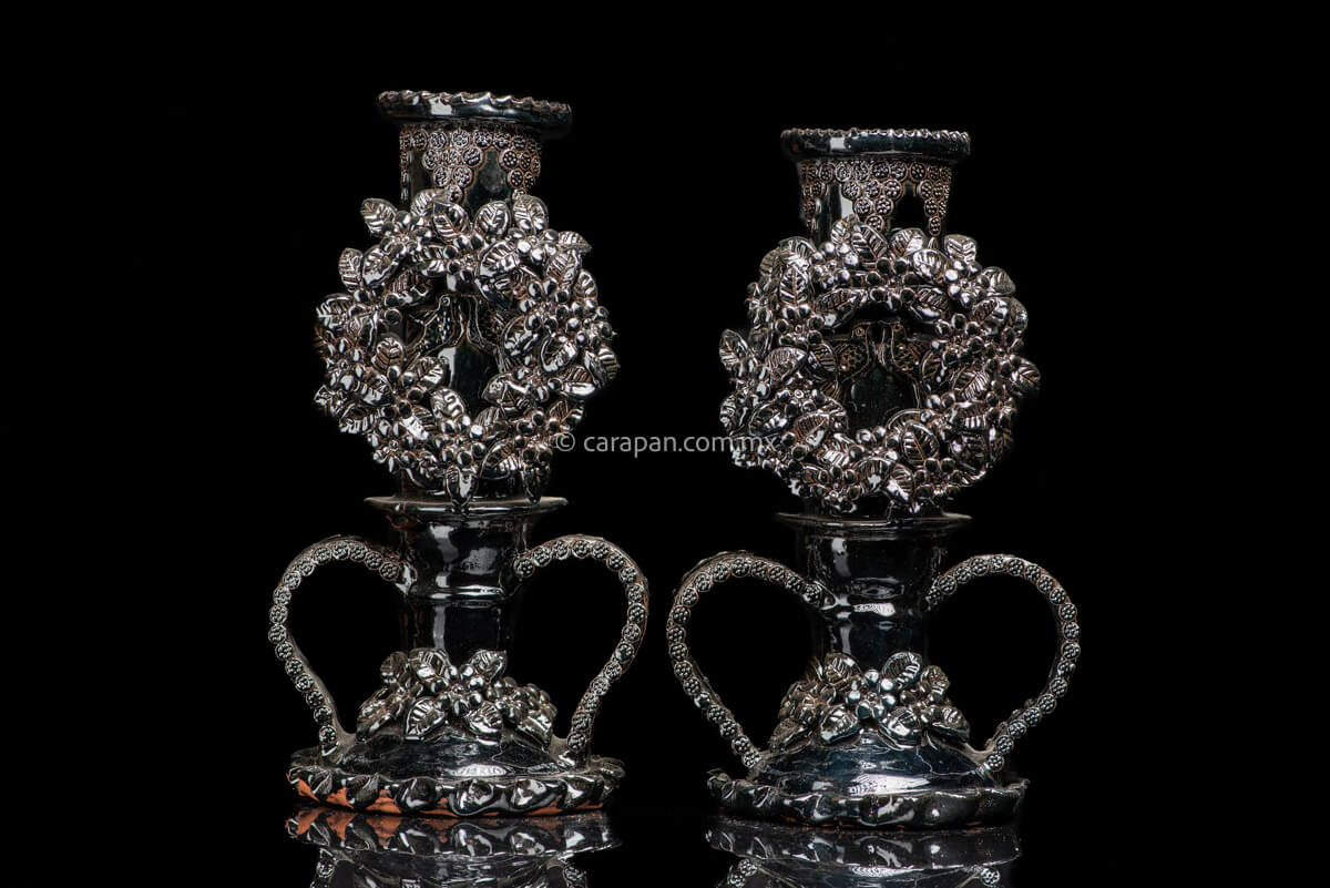 Set of 2 black glazed candlesticks. Each is decorated with a profuse pastillage crown of flowers. At the center of each crown is a couple of loving birds. Each candlestick has two  curved handles at the bottom. The contour of the handles is decorated with rosettes and the base of each candlestick is decorated with flowers and leafs.