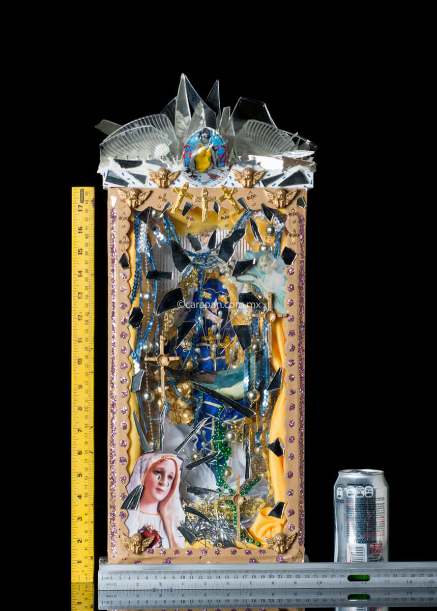 Kitsch Style Diorama by Mexican Artist Alfredo Torres with Relitious elements such as golden crosses, a saint in blue crafted with textile elements, mirror fragments, and clipping of religious image. The top of the diorama's box is decorated with broken glass and mirror fragments. At the bottom of the box a dolorosa virgin image clipping stands out. Both sides of the box have written prayers for protection.  Rulers