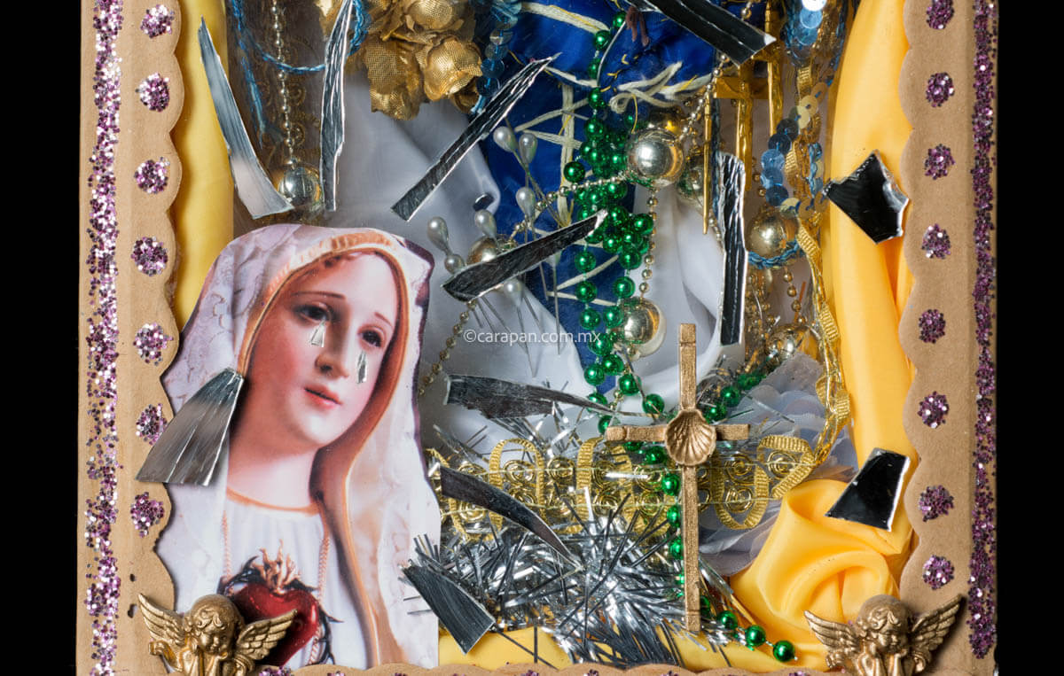 Kitsch Style Diorama by Mexican Artist Alfredo Torres with Relitious elements such as golden crosses, a saint in blue crafted with textile elements, mirror fragments, and clipping of religious image. The top of the diorama's box is decorated with broken glass and mirror fragments. At the bottom of the box a dolorosa virgin image clipping stands out. Both sides of the box have written prayers for protection. 
