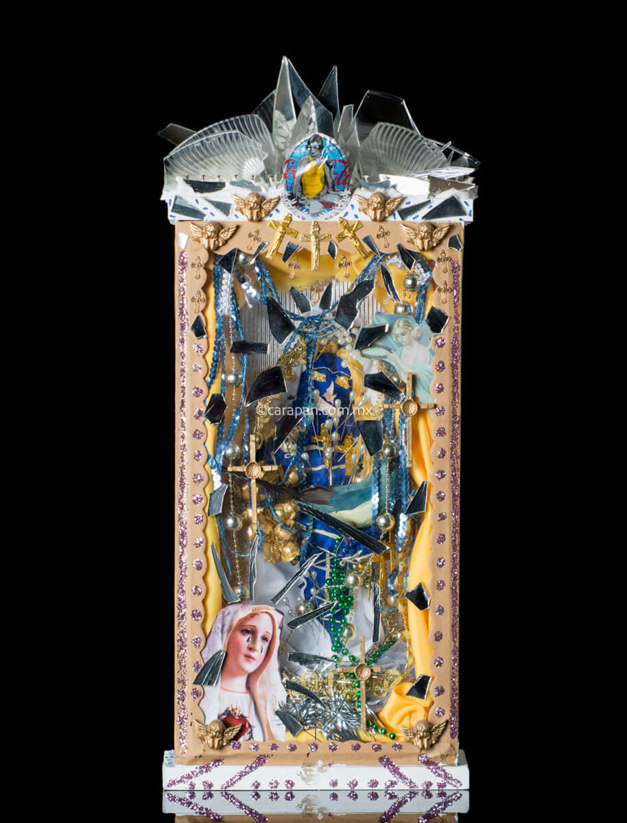 Kitsch Style Diorama by Mexican Artist Alfredo Torres with Relitious elements such as golden crosses, a saint in blue crafted with textile elements, mirror fragments, and clipping of religious image. The top of the diorama's box is decorated with broken glass and mirror fragments. At the bottom of the box a dolorosa virgin image clipping stands out. Both sides of the box have written prayers for protection. 