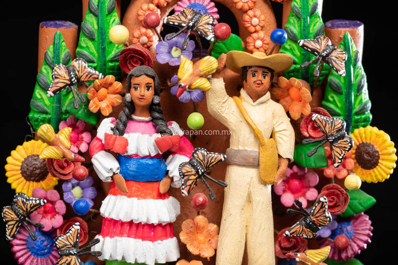 Small Mexican Clay tree of life with Mazahua indigenous couple at the center surrounded by flowers and butterflies. The man wears a traditional costume in beig, hat and a bag. The woman wears a traditional dress in pink, orange and green with a blue belt. 