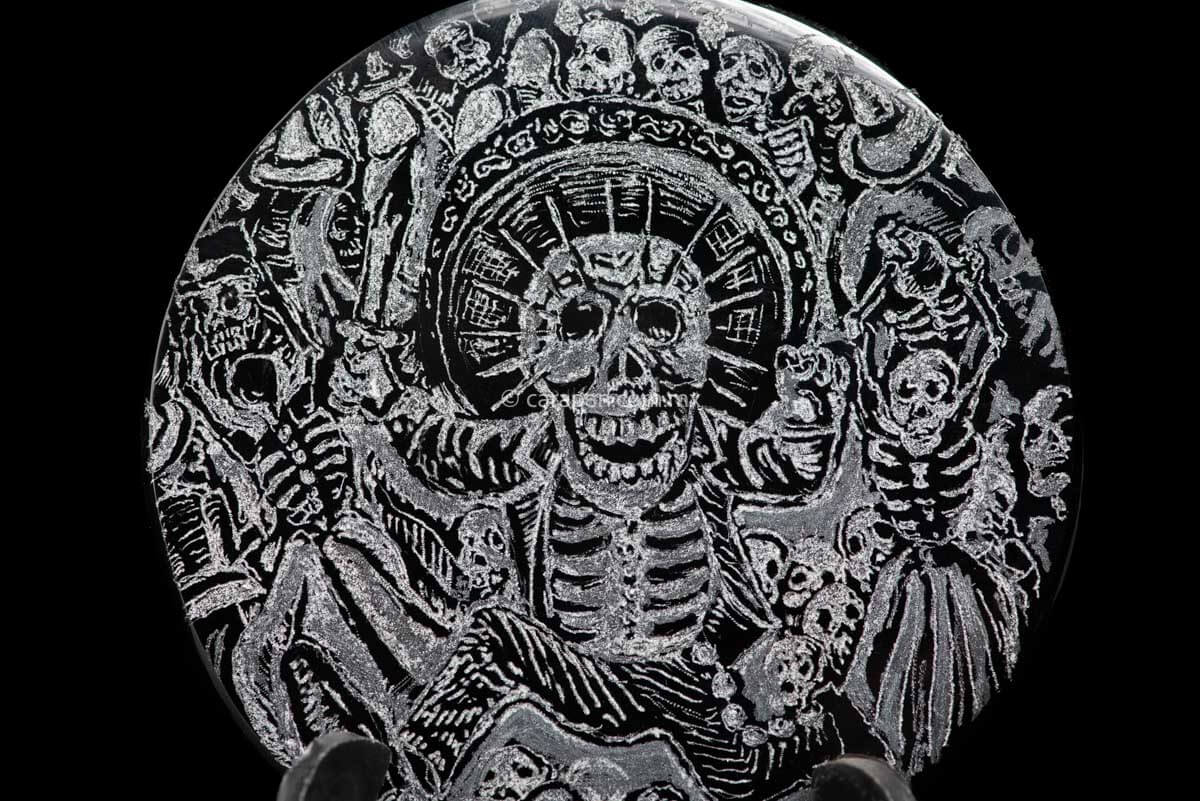 Obsidian disc with engraved skeleton wearing a charro costume, holding a knife on his right hand and running . Surrounded by skulls and skeleton. Based on Jose Guadalupe Posadas Work