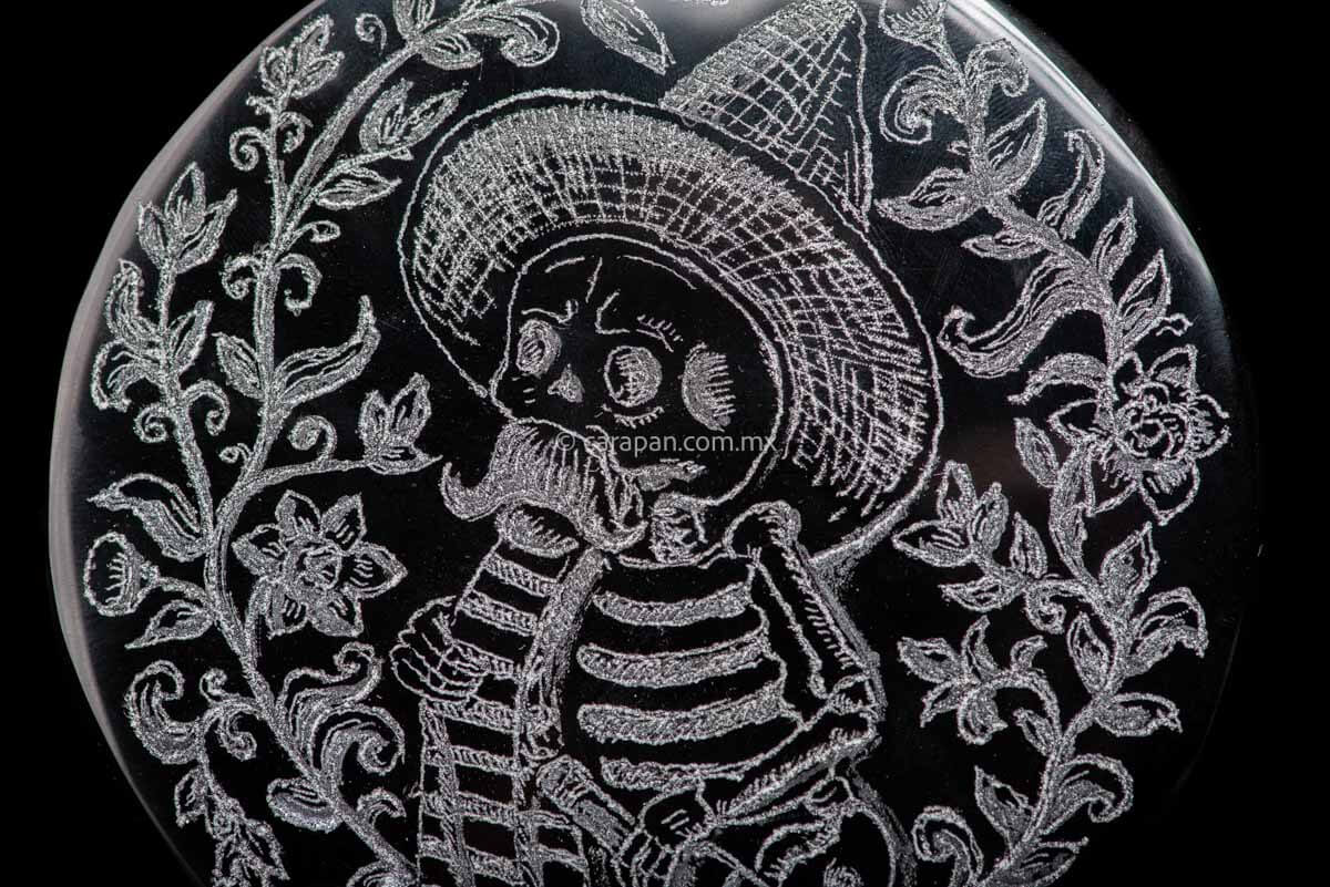 Skeleton  wearing a sombrero, a sarape and holding a bottle based upon a Jose Guadalupe Posadas drawing. The piece is framed by flowers and vegetal motifs.