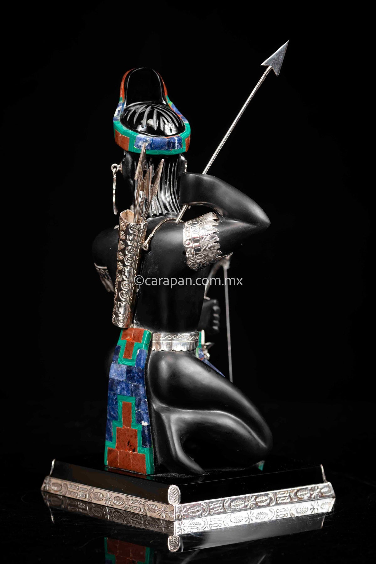 Obsidian Sculpture of Aztec Warrior with Bow & Arrows he is wearing two silver earrings, a silver necklace and two  silver sleeves with feathers