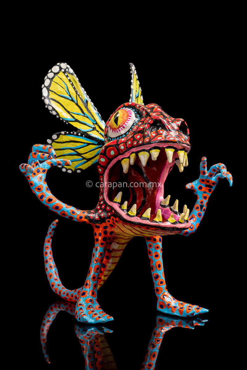 Paper Mache Alebrije with yellow butterfly wings, mouth open showing teeth in fierce expression, big eyes red body & blue legs & Arms decorated with red  patches and blue dots