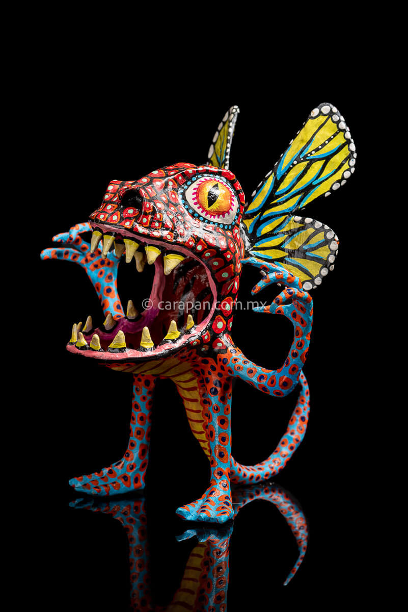 Paper Mache Alebrije with yellow butterfly wings, mouth open showing teeth in fierce expression, big eyes red body & blue legs & Arms decorated with red  patches and blue dots