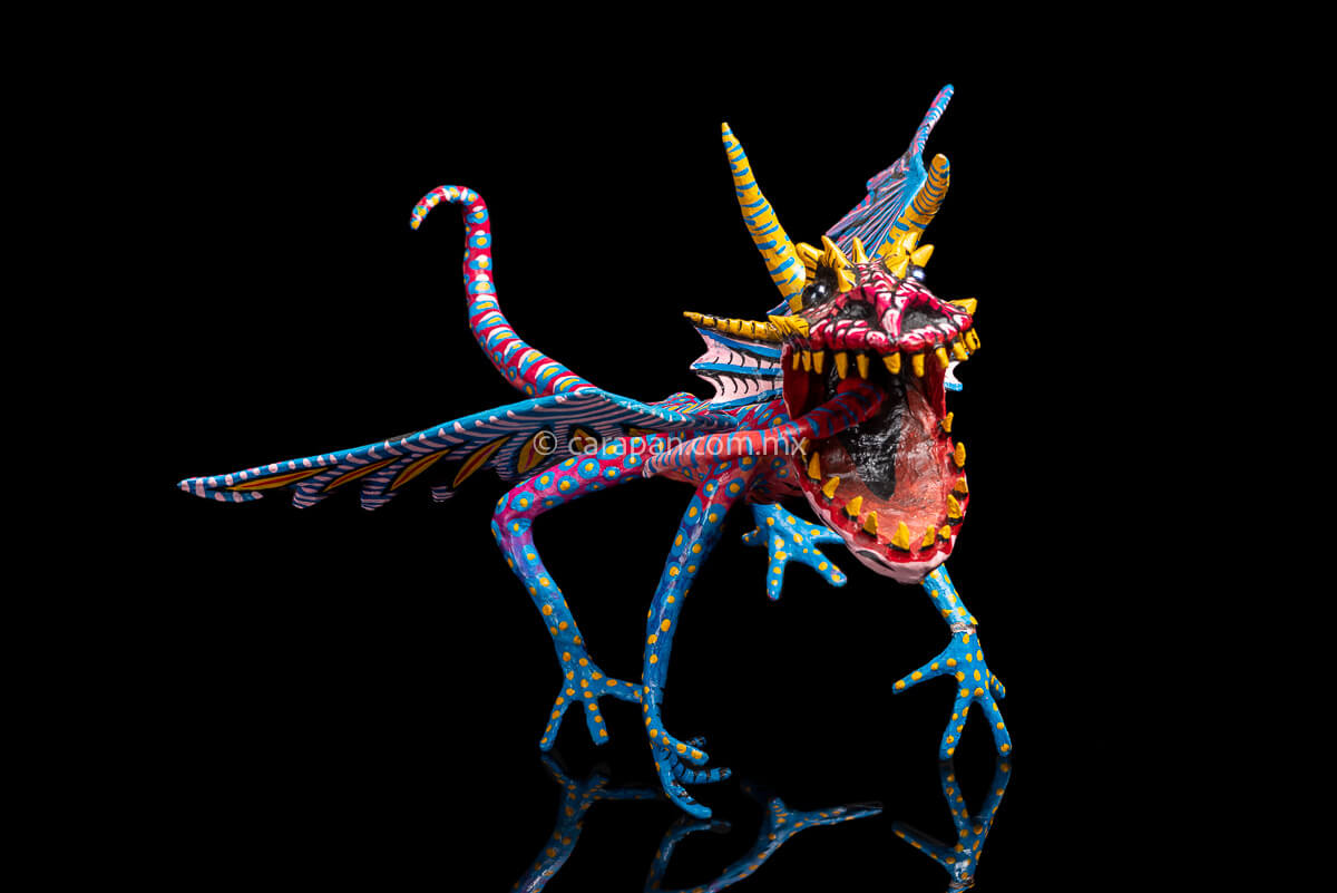 Mexican Paper Mache Alebrije  Dragon  style with fierce expression, two horns paionted in yellow with blue strokes, marble eyes, open mouth showing tootn, long red tongue with blue strokes, wings decorated in blue and pink tones on the outside and blue and white on the inside. Long tail decorated in red with light blue patches with yellow center. HAnds and legs decorated in blue with yellow dots. 