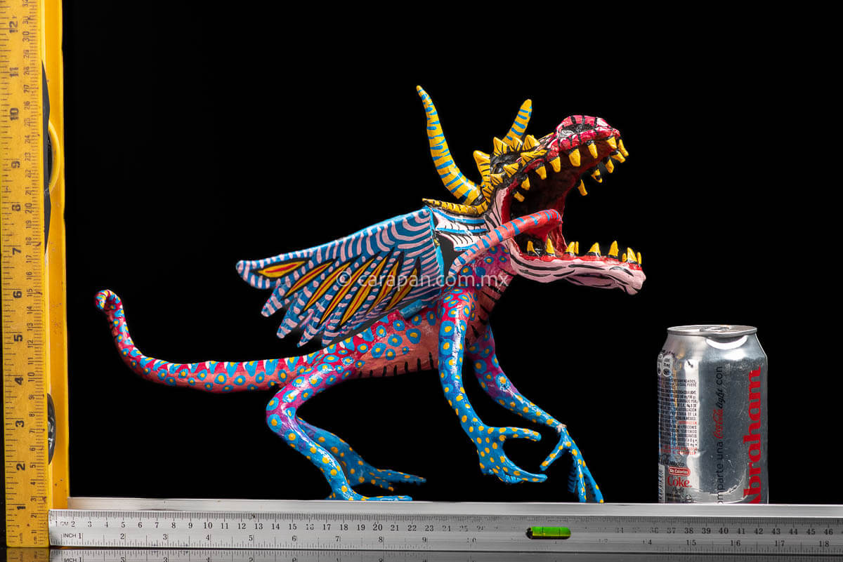 Mexican Paper Mache Alebrije  Dragon  style with fierce expression, two horns paionted in yellow with blue strokes, marble eyes, open mouth showing tootn, long red tongue with blue strokes, wings decorated in blue and pink tones on the outside and blue and white on the inside. Long tail decorated in red with light blue patches with yellow center. HAnds and legs decorated in blue with yellow dots. Rulers