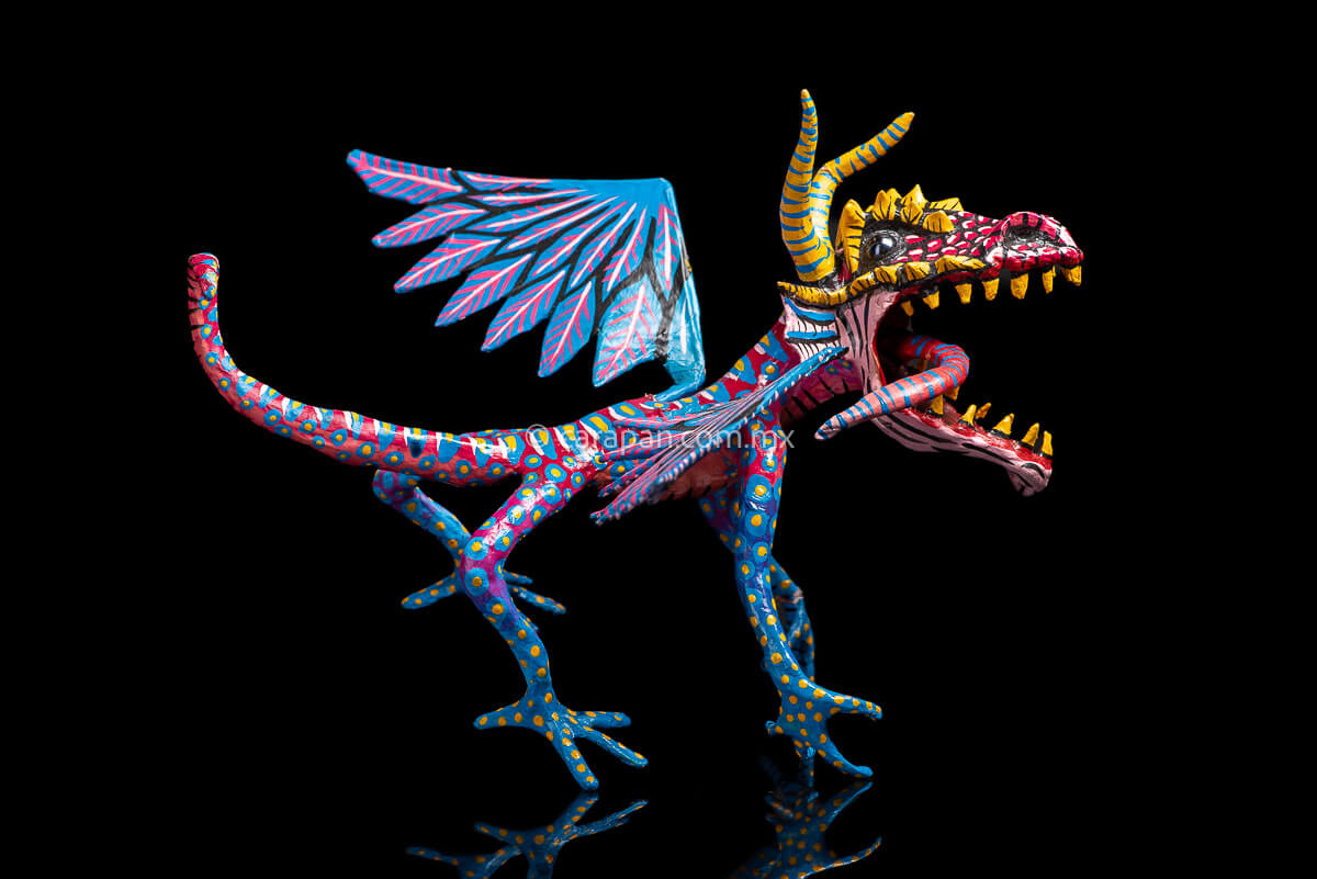 Mexican Paper Mache Alebrije  Dragon  style with fierce expression, two horns paionted in yellow with blue strokes, marble eyes, open mouth showing tootn, long red tongue with blue strokes, wings decorated in blue and pink tones on the outside and blue and white on the inside. Long tail decorated in red with light blue patches with yellow center. HAnds and legs decorated in blue with yellow dots. 