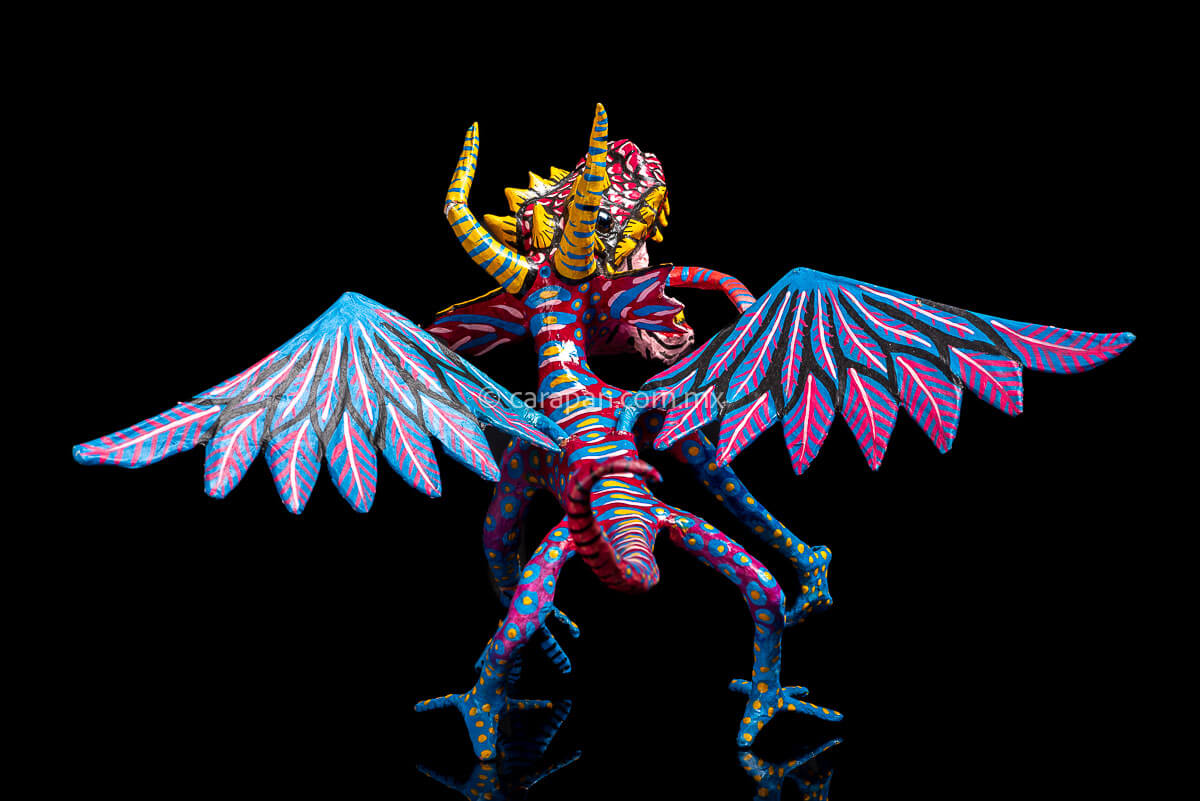 Mexican Paper Mache Alebrije  Dragon  style with fierce expression, two horns paionted in yellow with blue strokes, marble eyes, open mouth showing tootn, long red tongue with blue strokes, wings decorated in blue and pink tones on the outside and blue and white on the inside. Long tail decorated in red with light blue patches with yellow center. HAnds and legs decorated in blue with yellow dots.  back